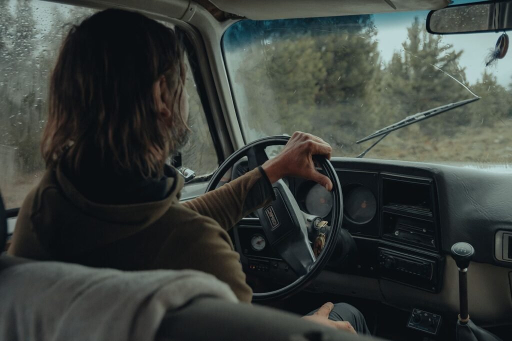 A person drives a vintage car through a wooded area on a rainy day, viewed from behind.