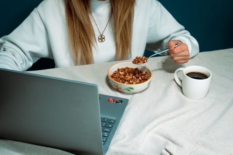 Woman enjoying cereal breakfast while browsing on laptop, featuring coffee and casual work setup.