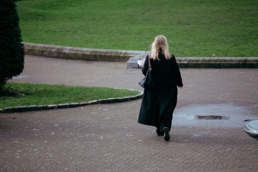 A woman in a black coat walks alone in a peaceful Dublin park on a rainy day.