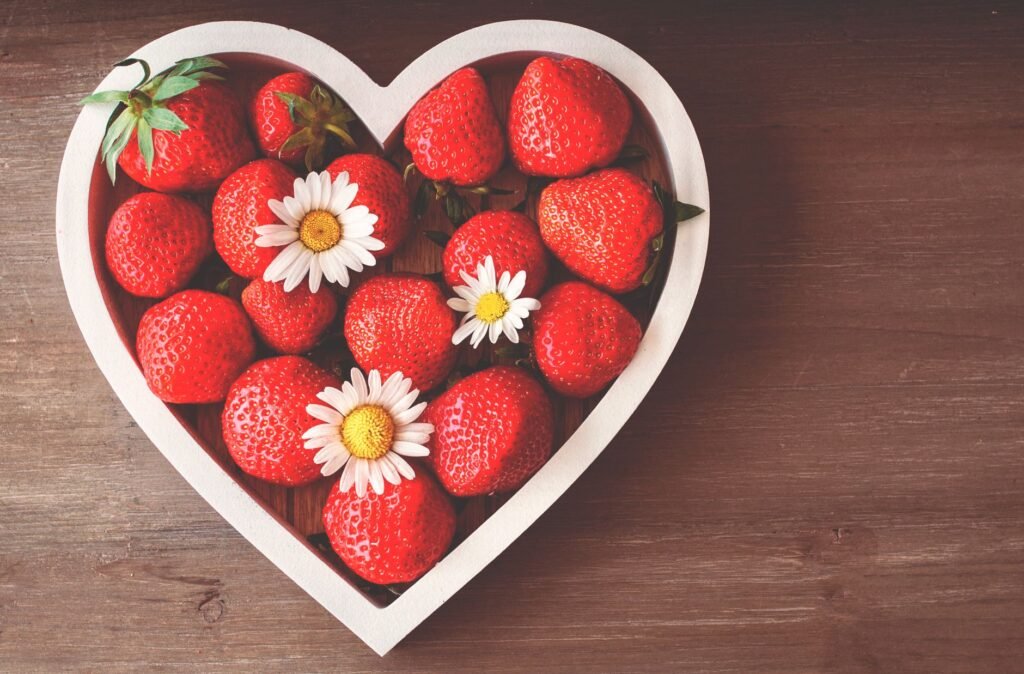 A heart-shaped bowl filled with fresh strawberries and daisies on a wooden surface, captured from above.