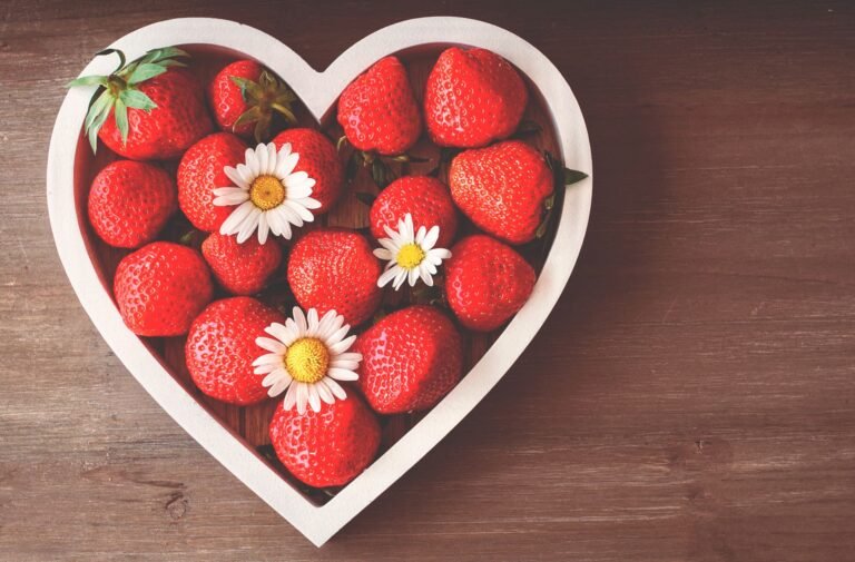 A heart-shaped bowl filled with fresh strawberries and daisies on a wooden surface, captured from above.