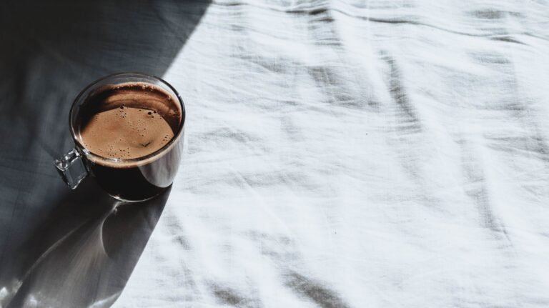 Glass cup of coffee casting shadows on a bright white sheet in natural sunlight.