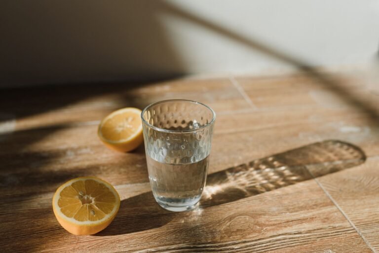 Clear glass of water with sliced lemons on a wooden surface in natural sunlight.