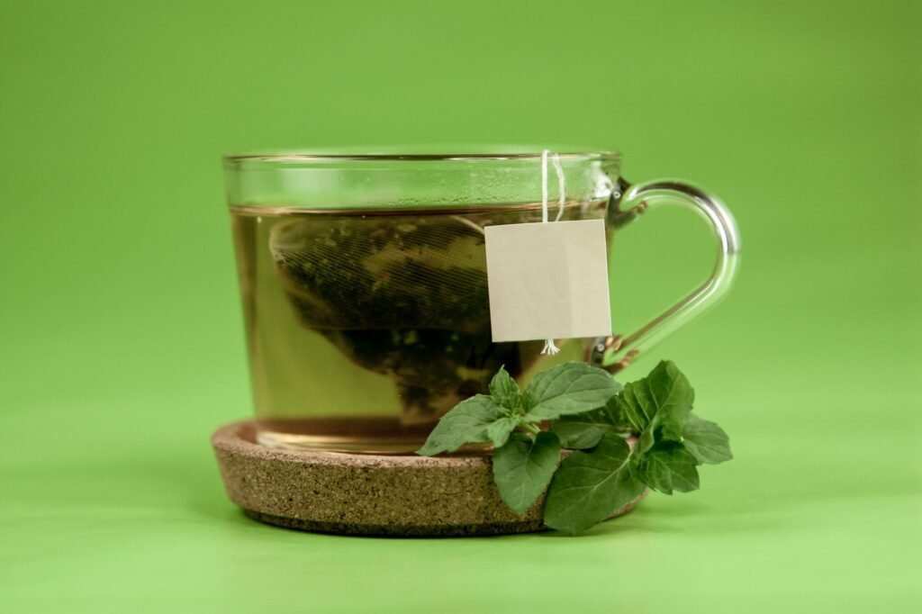 Transparent glass of hot green tea placed on ceramic cup holder with mint leaves on light green surface