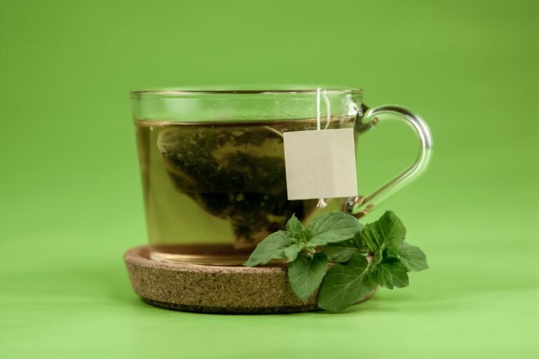 Transparent glass of hot green tea placed on ceramic cup holder with mint leaves on light green surface