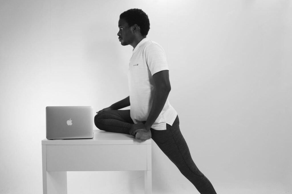 Black and white image of a man doing a stretch beside a laptop in a home office.