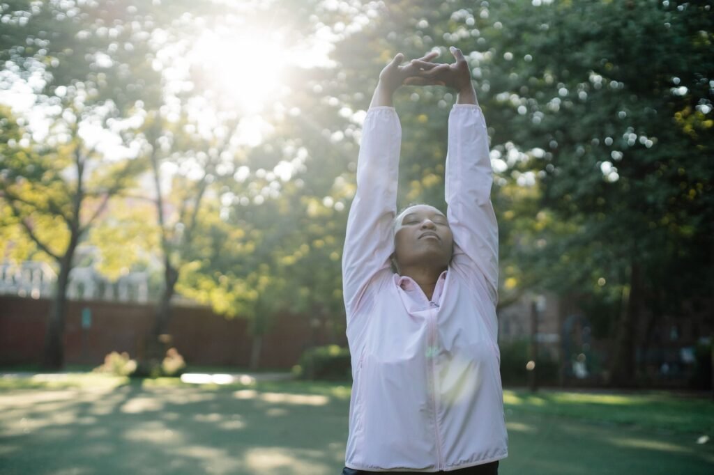 Black woman in pink jacket stretching in sunlit park, embracing a healthy lifestyle.