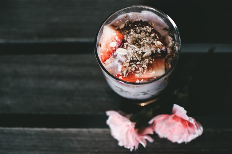 Glass of strawberry yogurt parfait with granola on a wooden table, garnished with pink flowers.