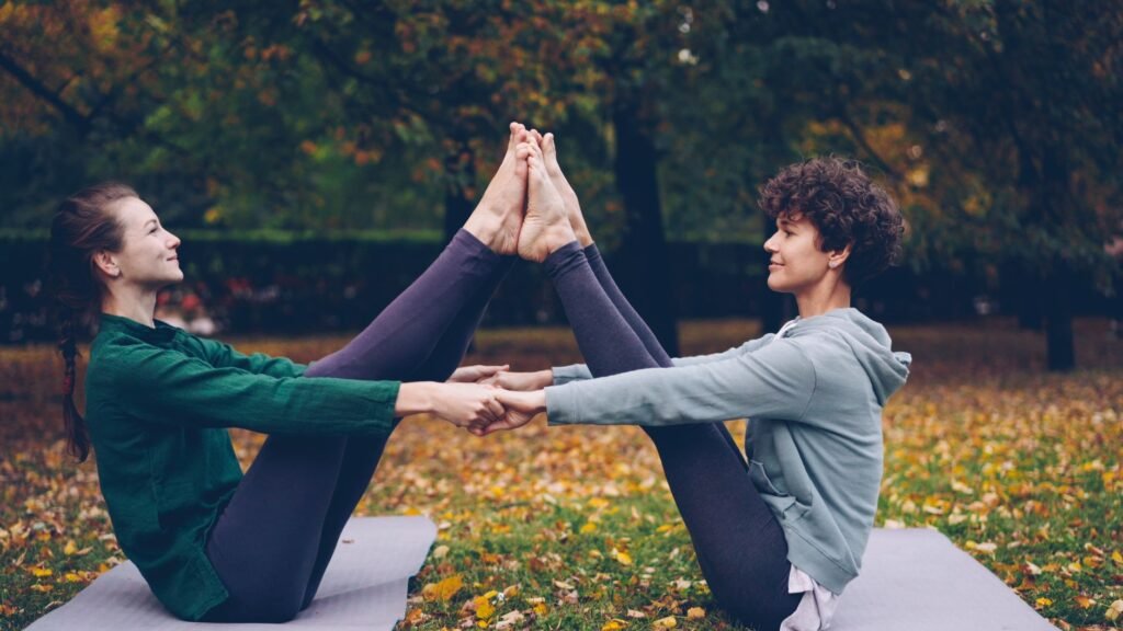 Two women practicing partner yoga outdoors on a fall day, surrounded by colorful leaves.
