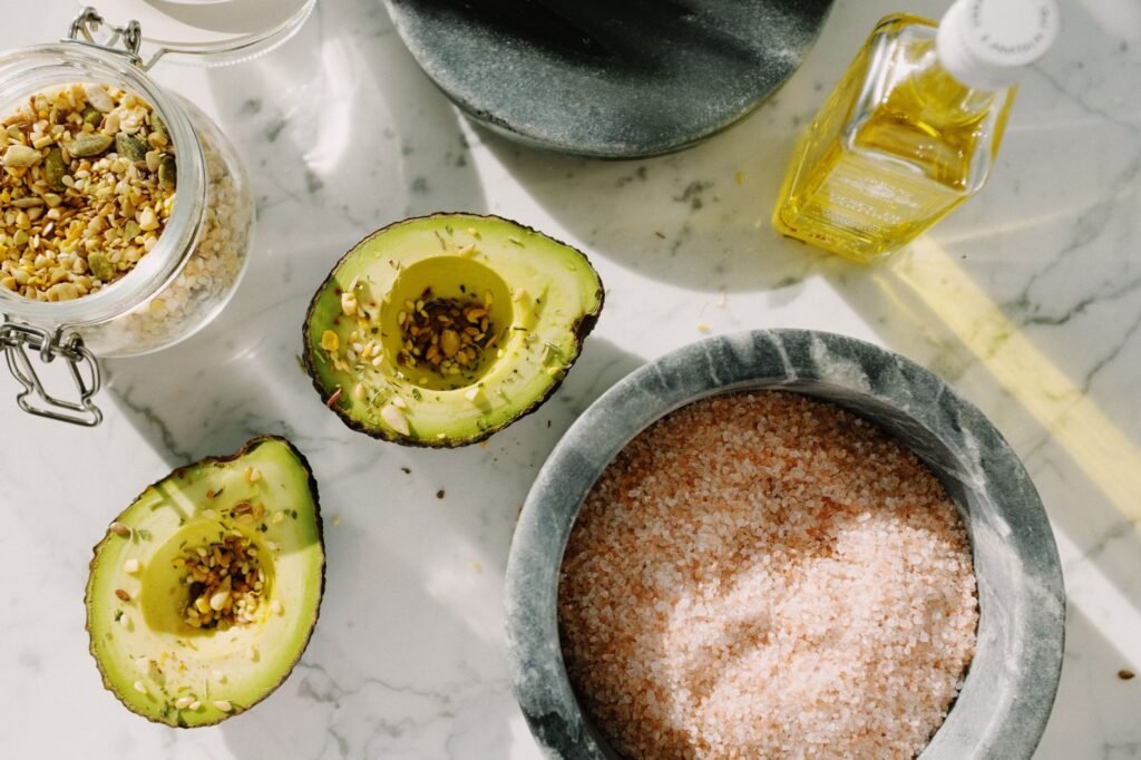 From above composition of fresh avocado pieces sprinkled with seeds from glass pot of seed mixture placed near bowl of natural olive oil and pink salt on white marble table