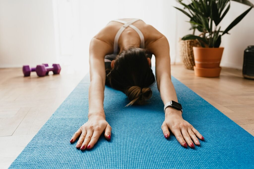 Full body faceless fit female wearing smartwatch stretching body in Child Pose on blue mat placed near plants and dumbbells in light living room