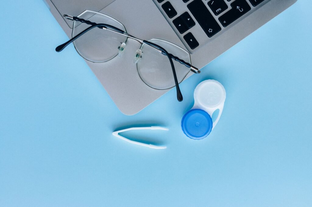 Flat lay of eyeglasses, contact lens case, and laptop on a blue surface.