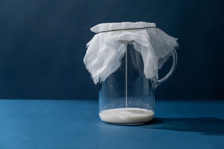 Glass pitcher with cheesecloth straining milk; minimalist still life on blue background.