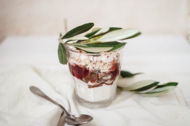 Tasty yogurt with chocolate sauce and coconut flakes under olive sprig in glass on crumpled textile
