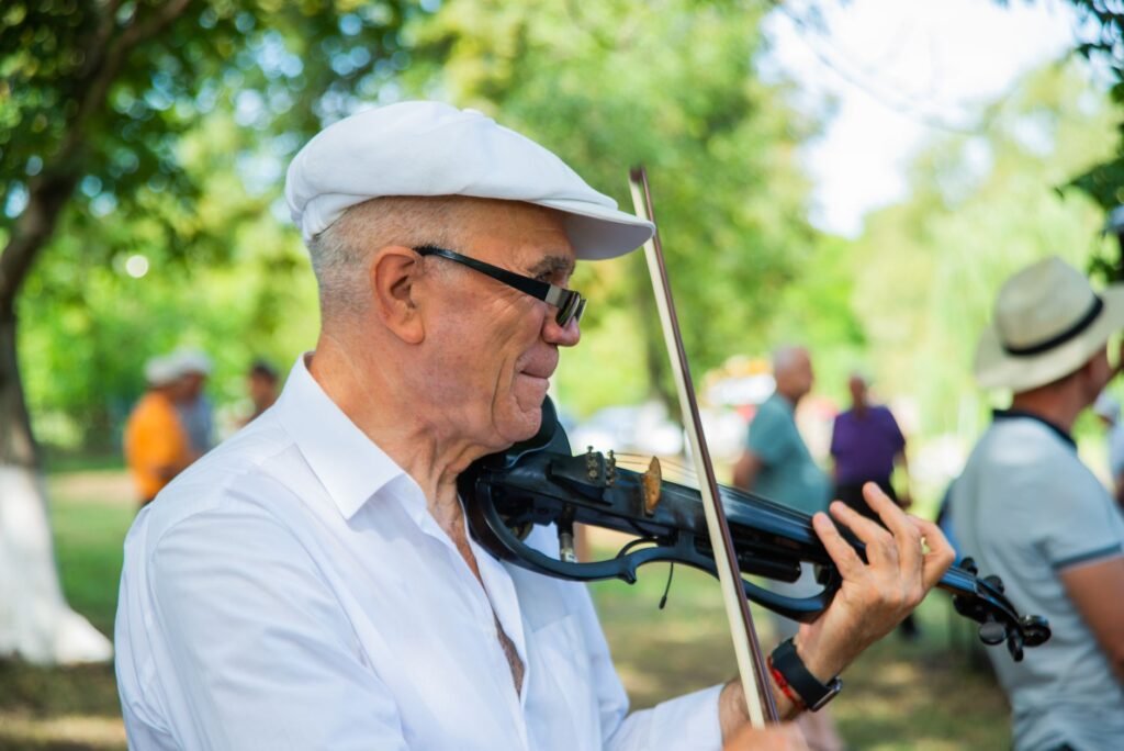 An elderly man joyfully plays the violin in a sunny park surrounded by people.
