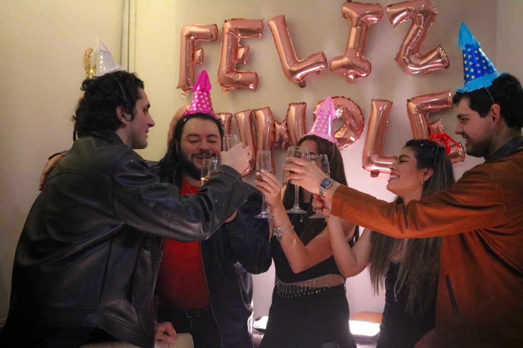 Five friends toasting with champagne at a birthday party, wearing colorful hats.