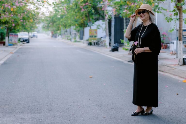 Senior woman in black dress and sun hat strolling on a peaceful tree-lined street.