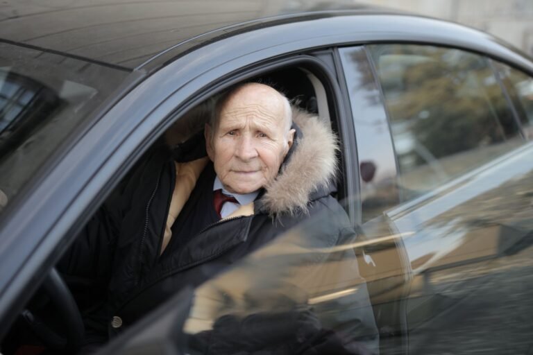 Senior man driving a car, wearing a winter coat, captured through the vehicle window.