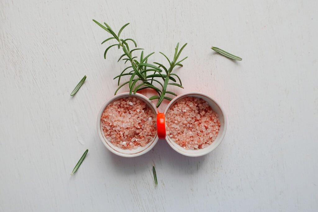 Top view of pink Himalayan salt and fresh rosemary in bowls on white table.