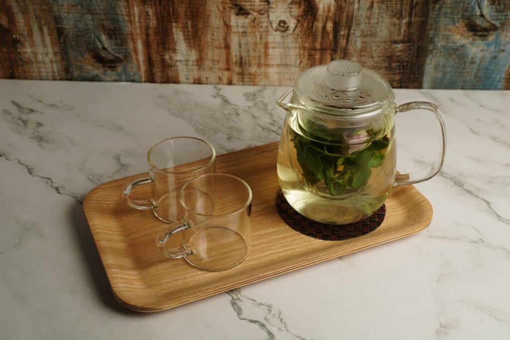 Glass teapot with fresh mint tea and glass mugs on a wooden tray, creating a cozy indoor setting.