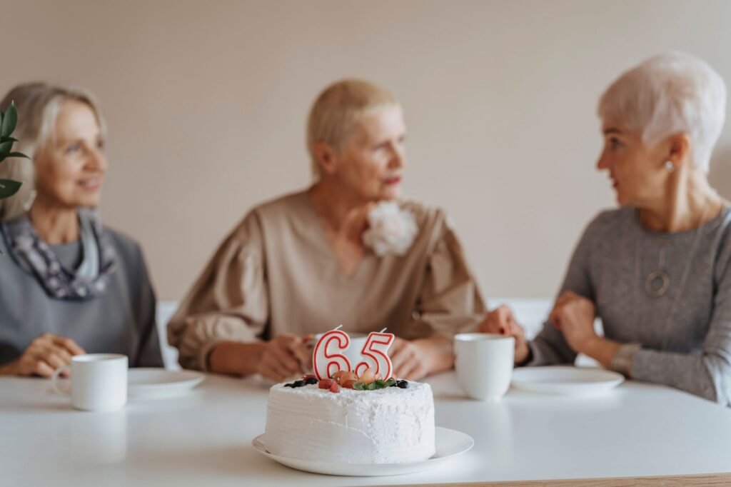 Three senior women celebrate a 65th birthday with cake and coffee indoors.