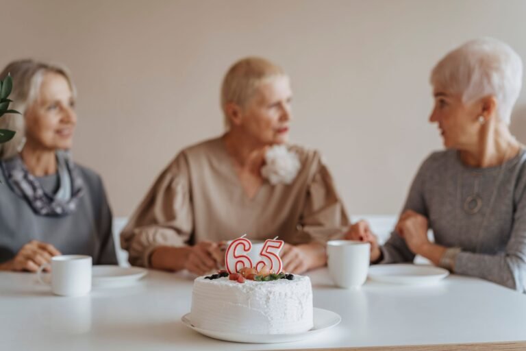 Three senior women celebrate a 65th birthday with cake and coffee indoors.