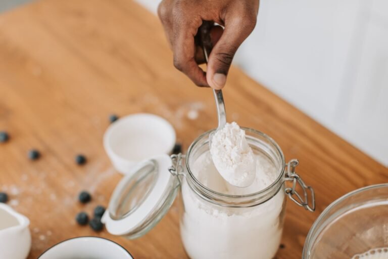 A person scooping flour from a glass jar on a wooden table surrounded by blueberries.