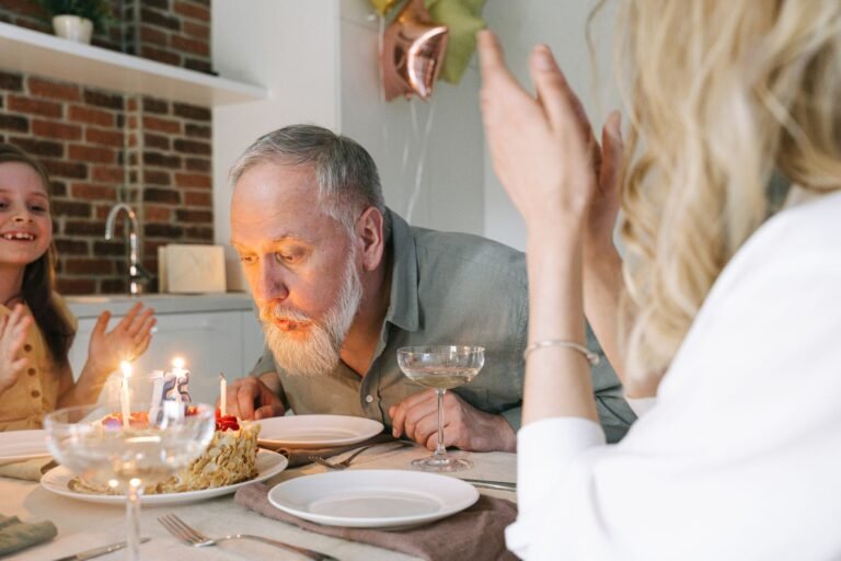 A senior man celebrates his birthday by blowing out candles on a cake in a cozy indoor setting.