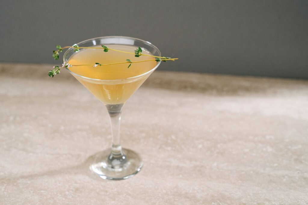 A stylish yellow cocktail garnished with fresh herbs in a martini glass against a neutral backdrop.