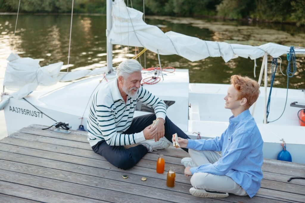 Senior man and young boy sharing snacks and laughter by a docked sailboat.