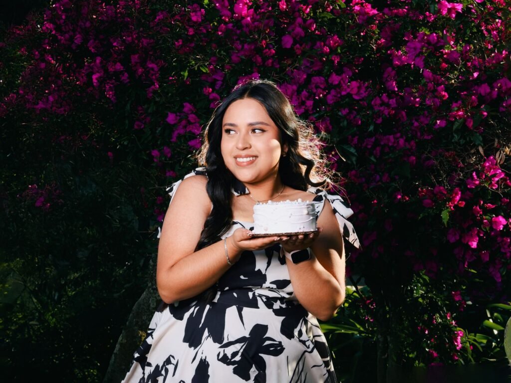 Smiling woman holding a cake in a floral outdoor setting, joyful and vibrant.